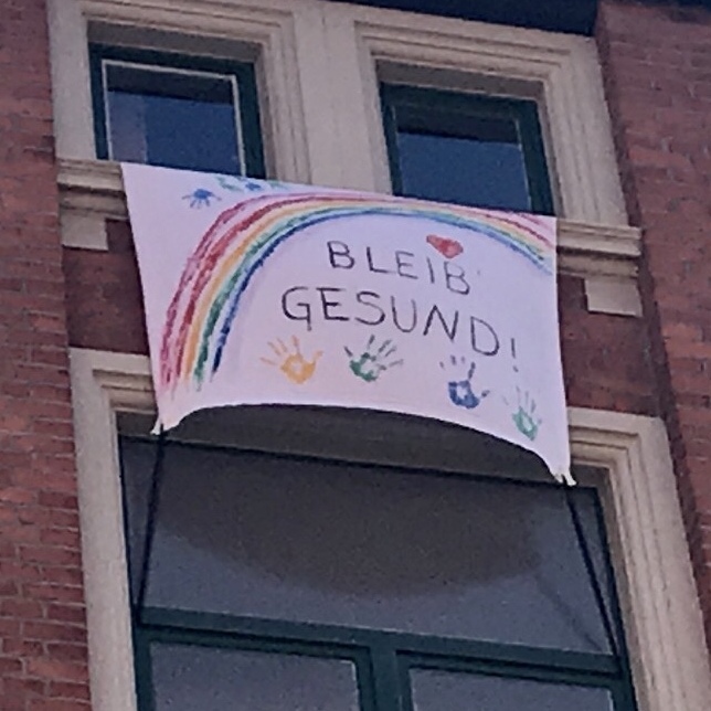 A bed sheet painted with a rainbow hangs on a house wall. It bears the inscription: stay healthy!